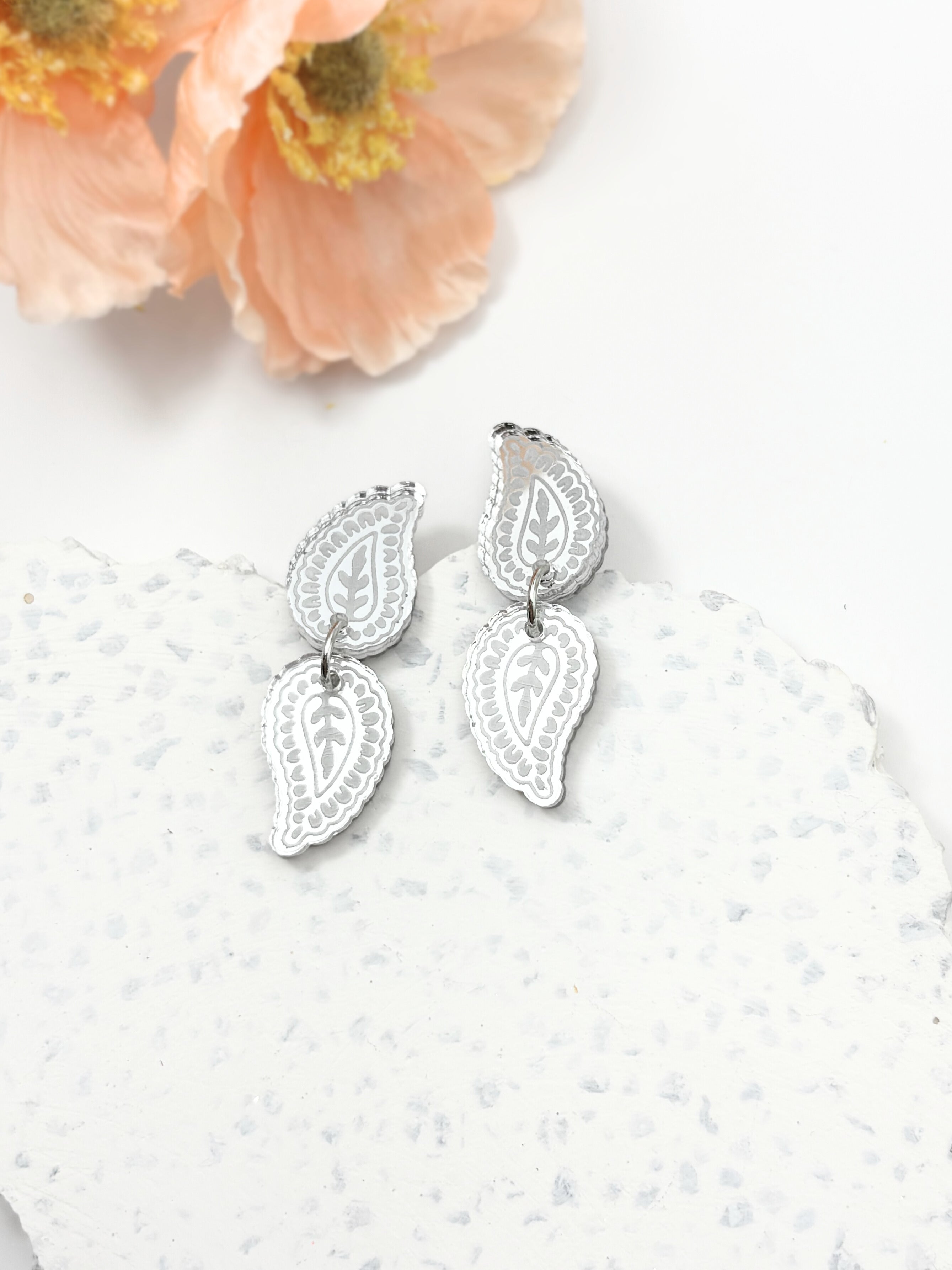 Silver earrings on a textured white surface with a peach flower in the corner.