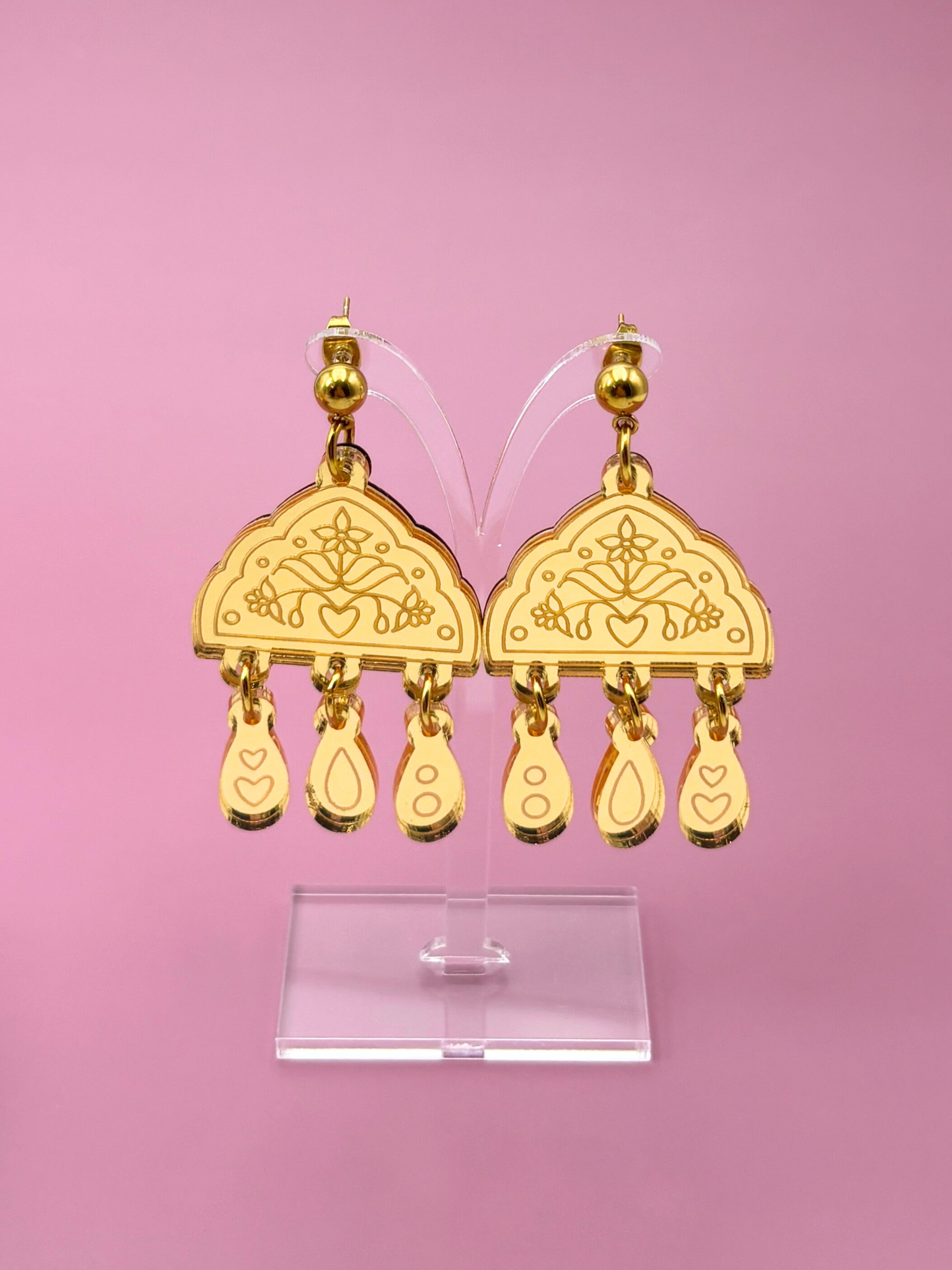 Gold earrings on a clear stand against a pink background