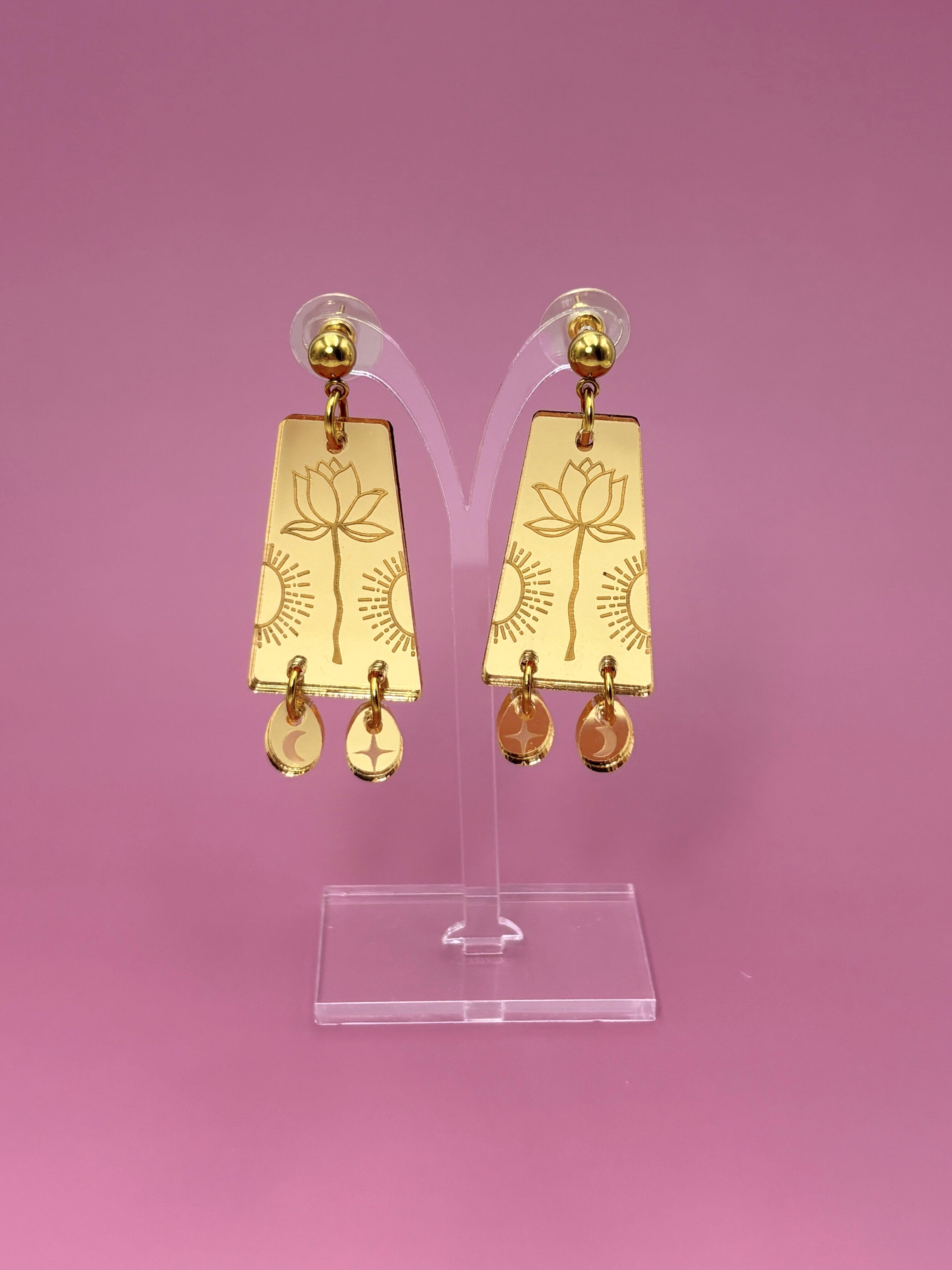 Gold earrings with floral design on a clear stand against a pink background