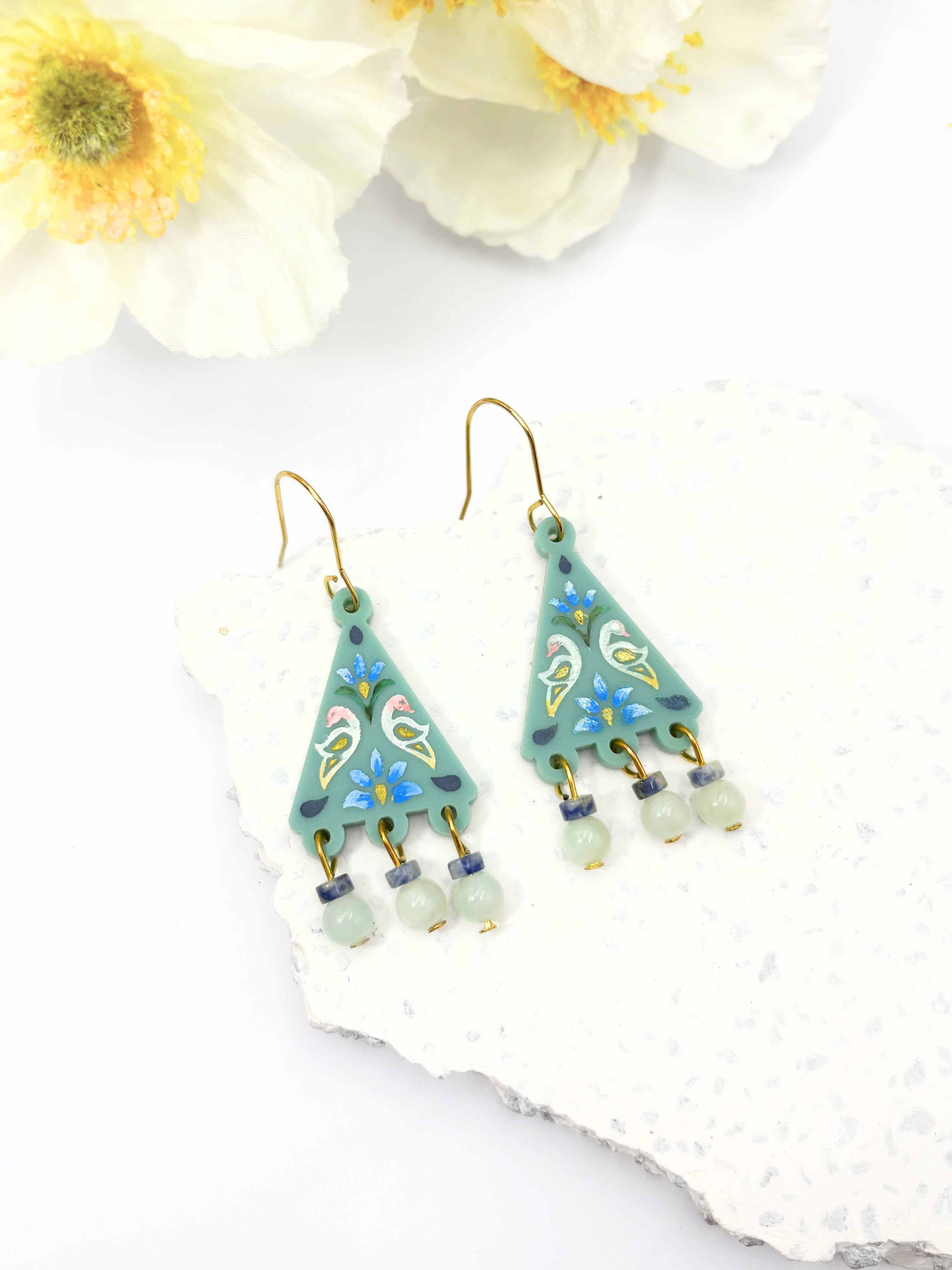 Pair of green triangle-shaped earrings with bead decorations on a white surface with flowers.