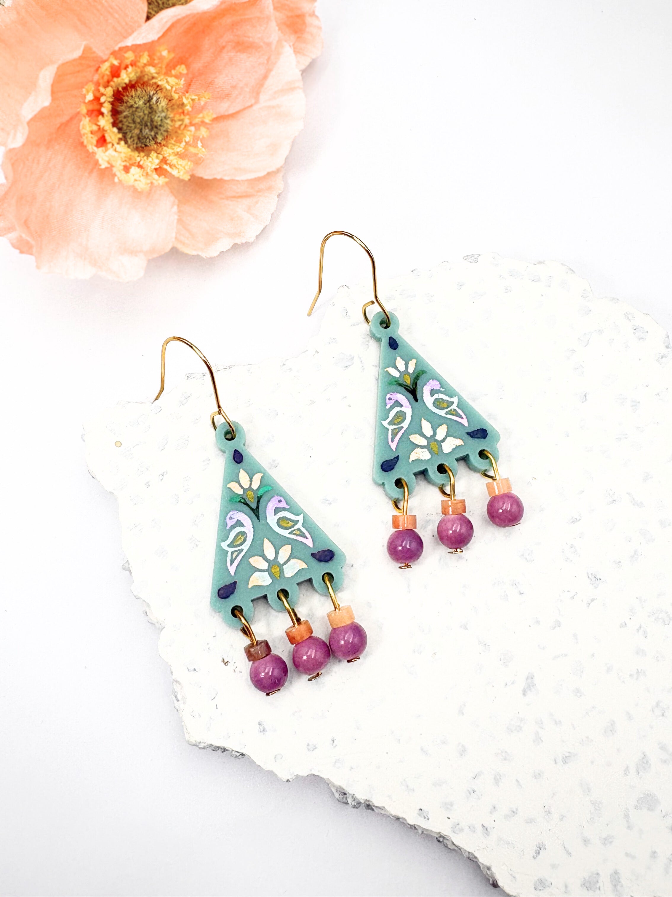 Teal geometric earrings with pink beads on a white surface with a flower.