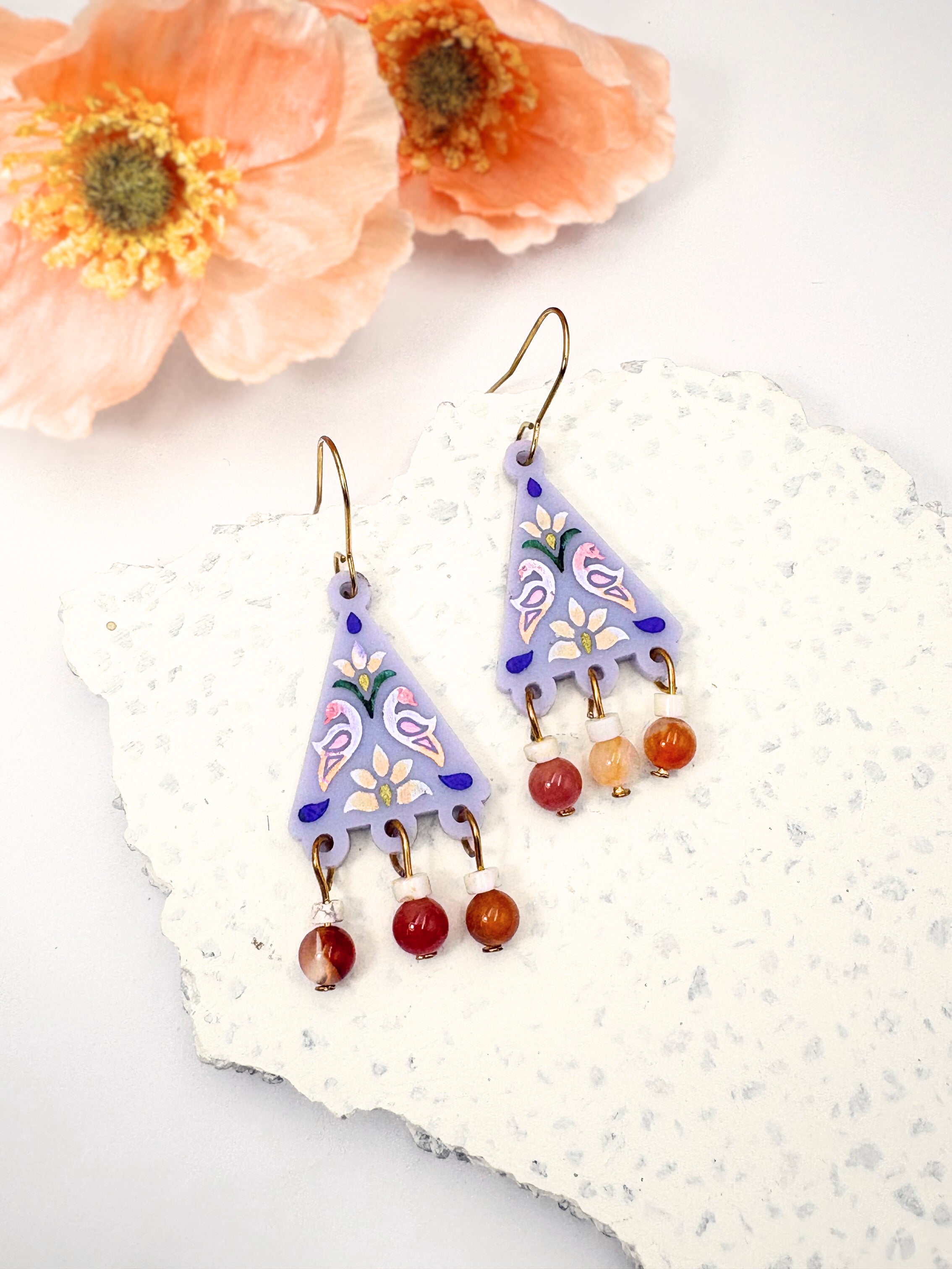 Colorful triangle-shaped earrings with red beads on a textured white surface with flowers.