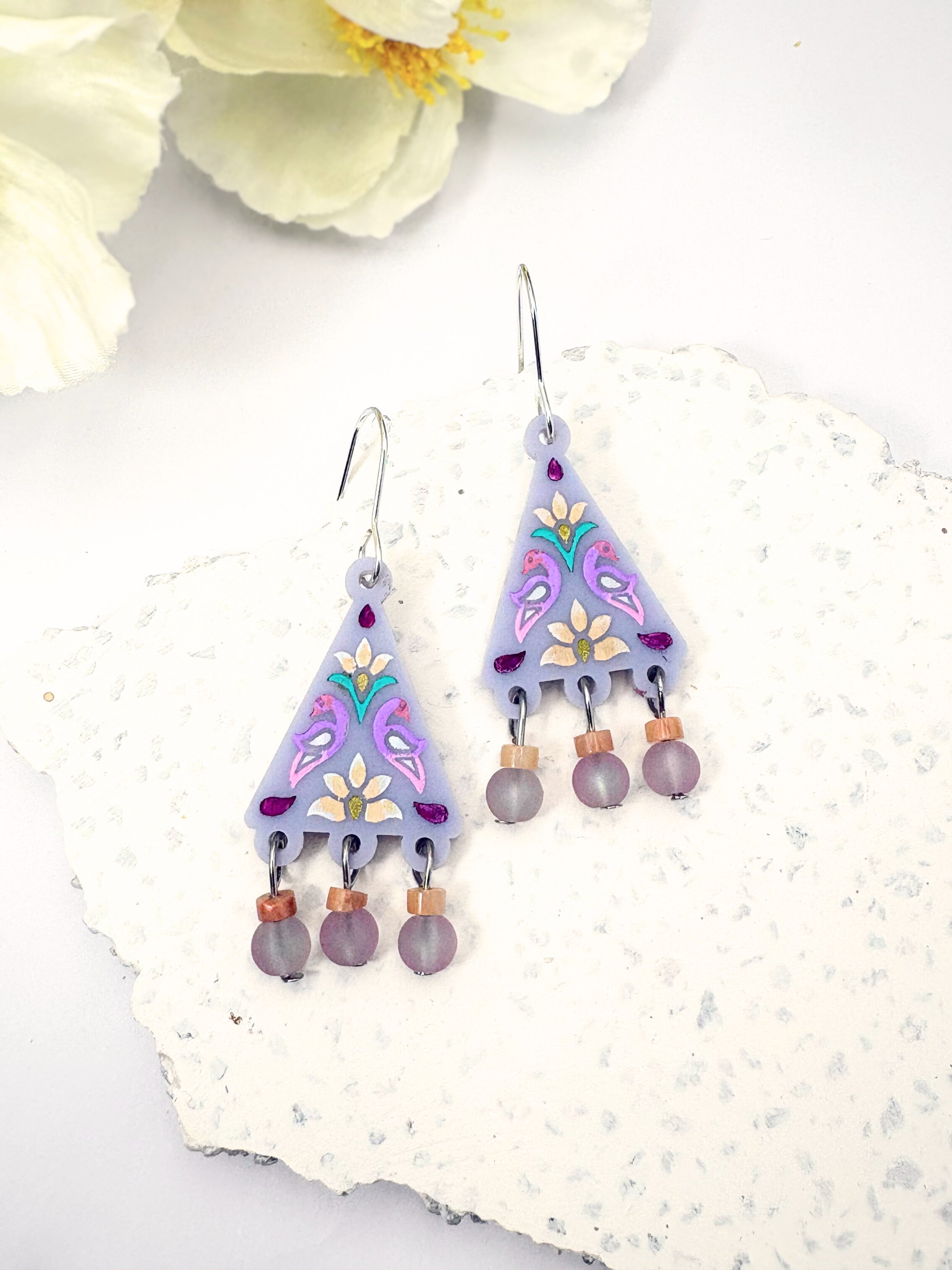Pair of purple floral earrings on a white surface with flowers in the background
