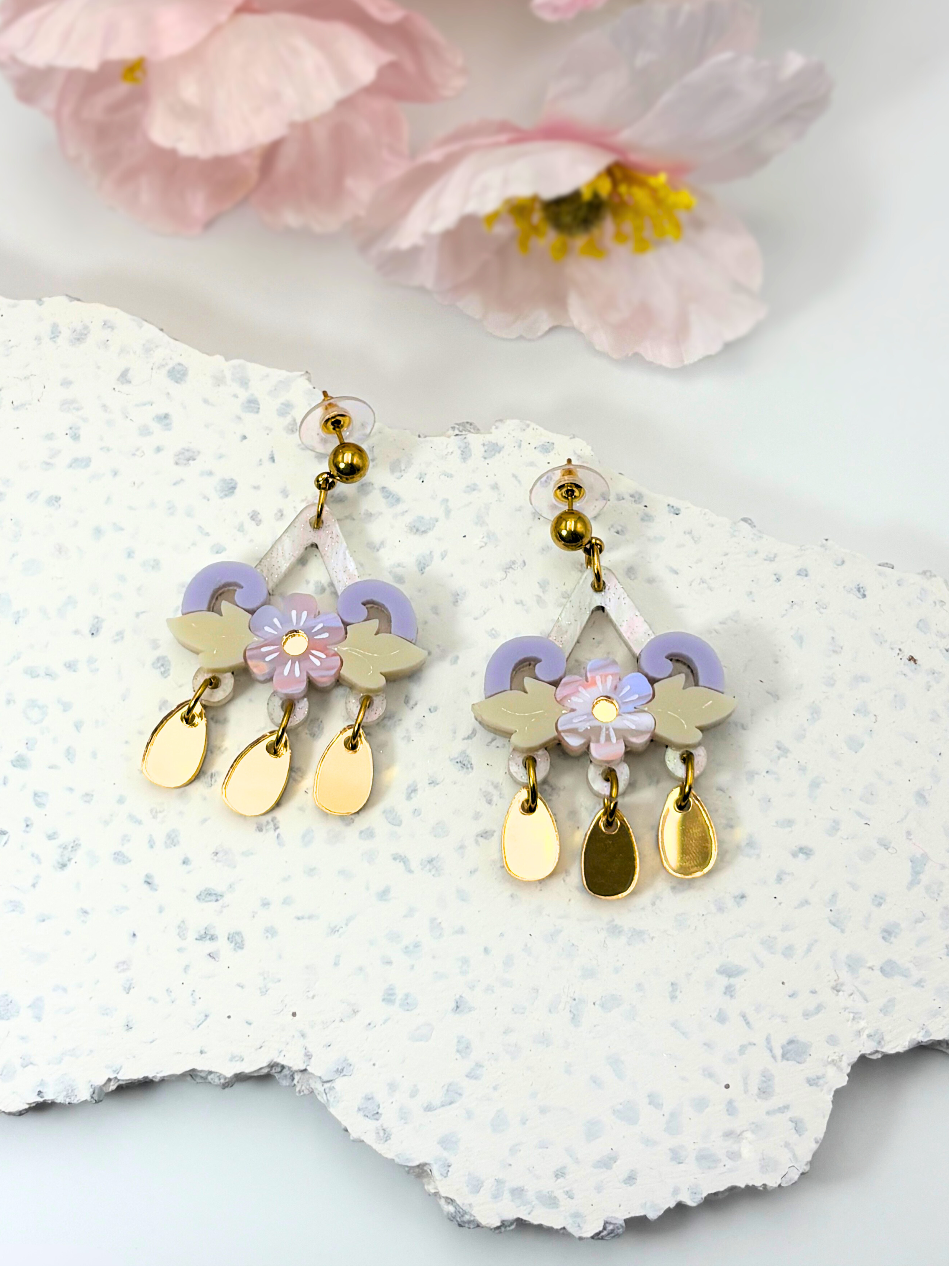 Chandelier earrings with floral and leaf designs on a textured white surface with pink flowers in the background.