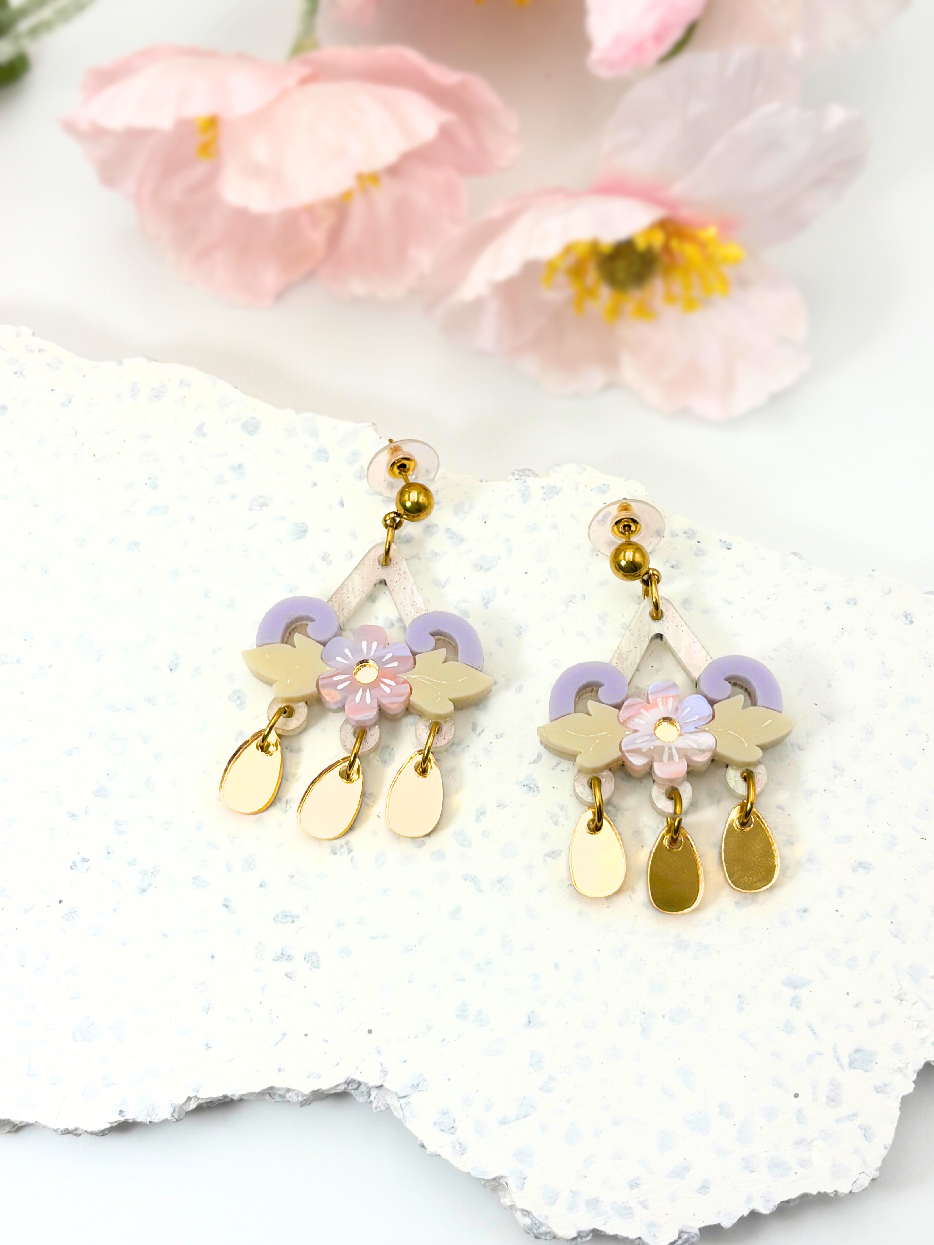 Pair of gold and pastel earrings on a white stone with pink flowers in the background