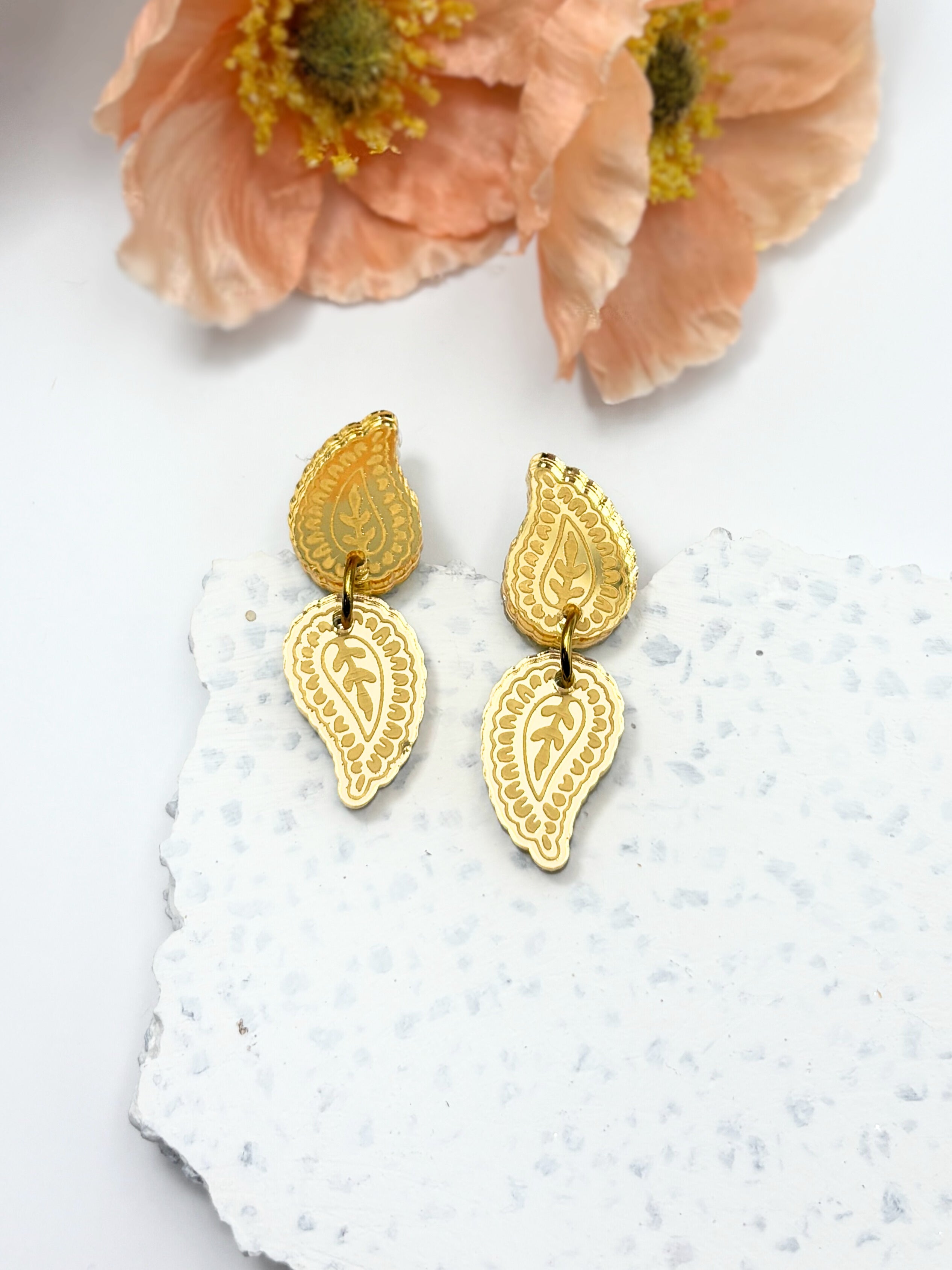 Gold earrings with intricate designs on a textured white surface with peach flowers in the background.