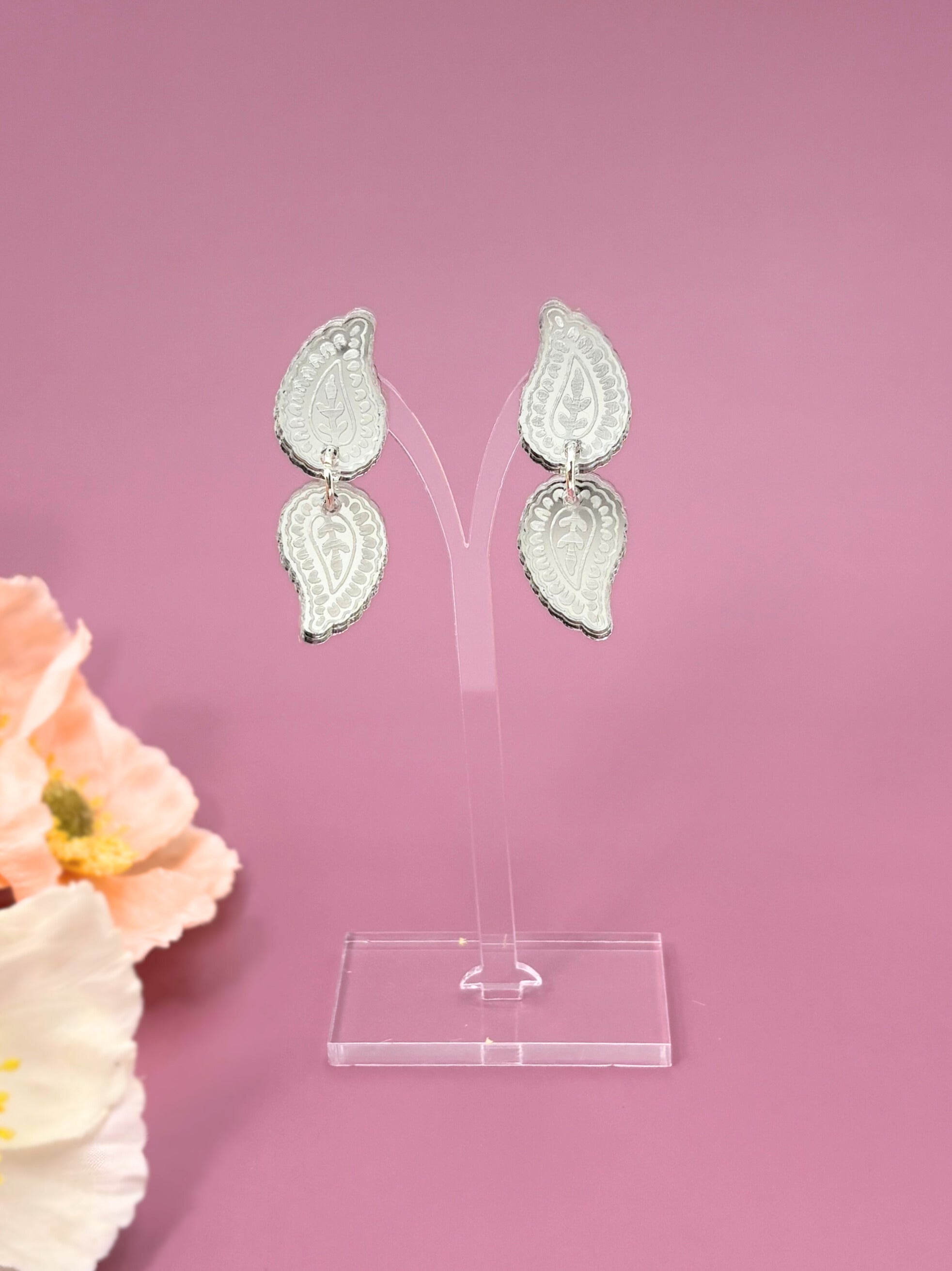 Silver leaf-shaped earrings on a clear stand against a pink background