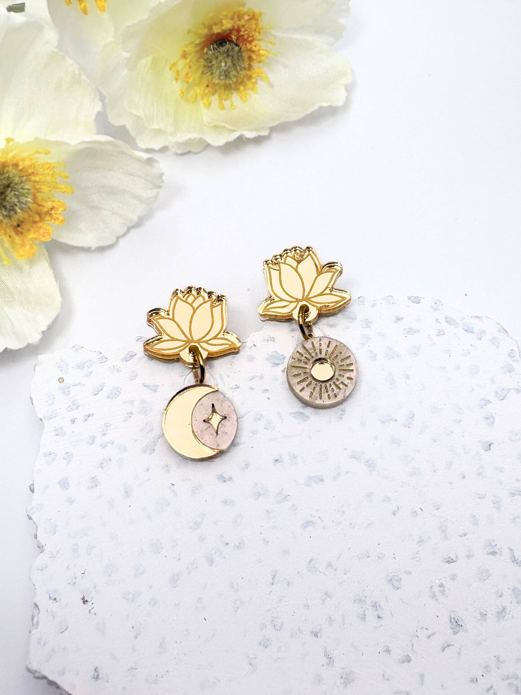 Gold lotus flower earrings with pink accents on a textured white surface with flowers.