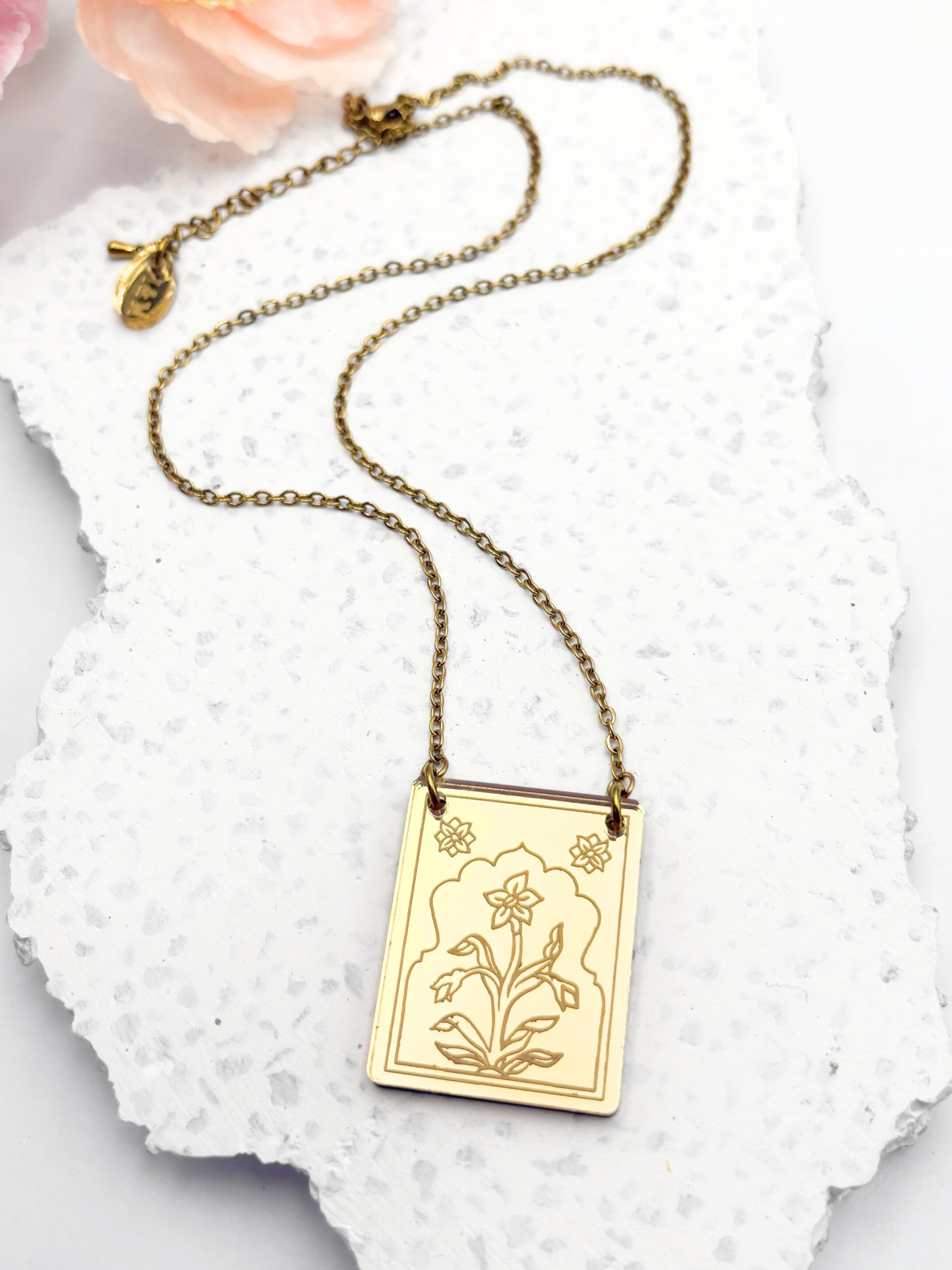 Gold necklace with a floral pendant on a textured white surface with flowers in the background