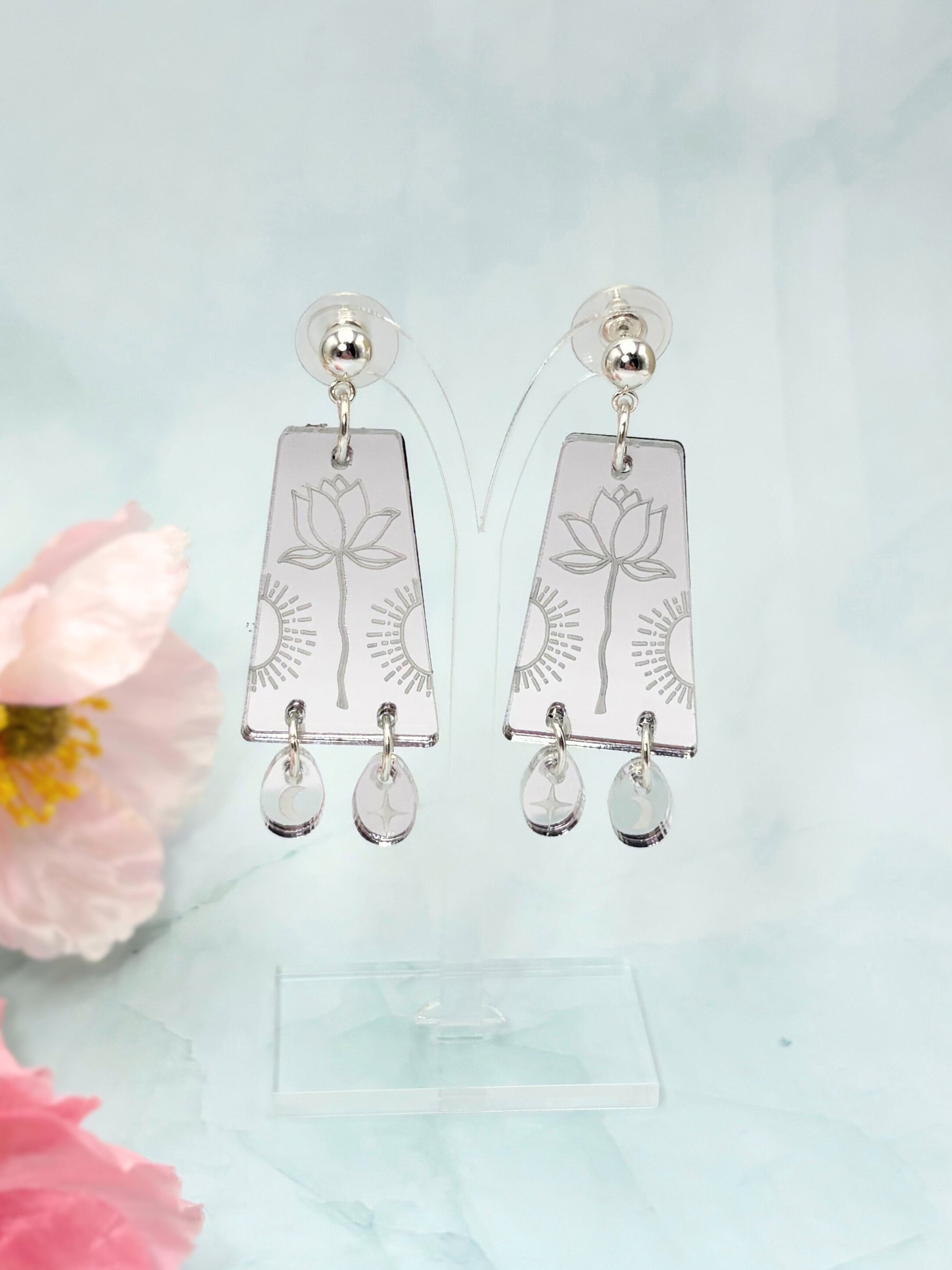 Silver earrings with floral designs and clear stones on a light blue background with pink flowers.