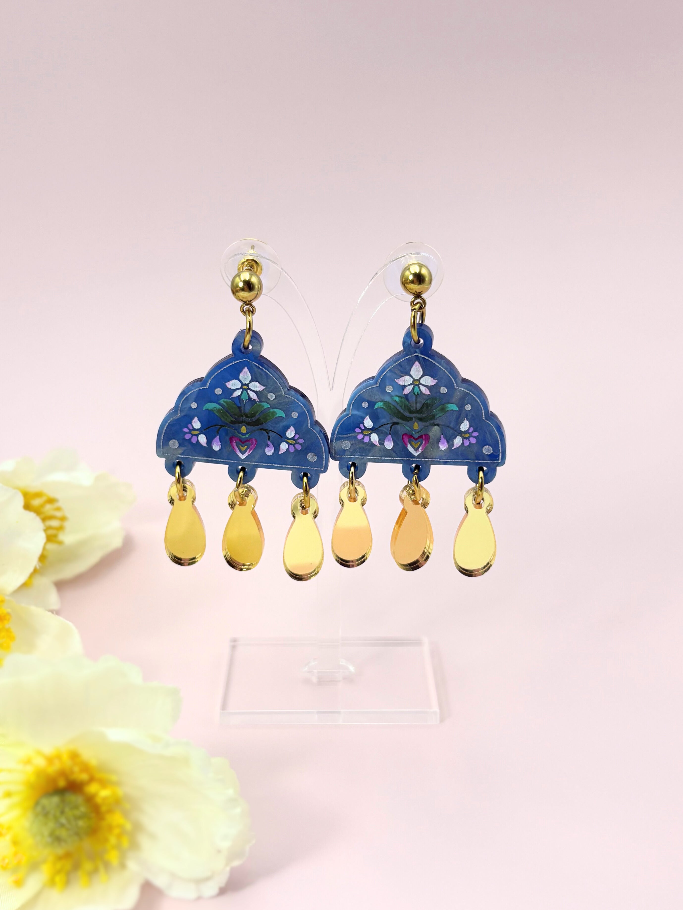 Decorative earrings with gold teardrop shapes and gold accents on a light pink background.