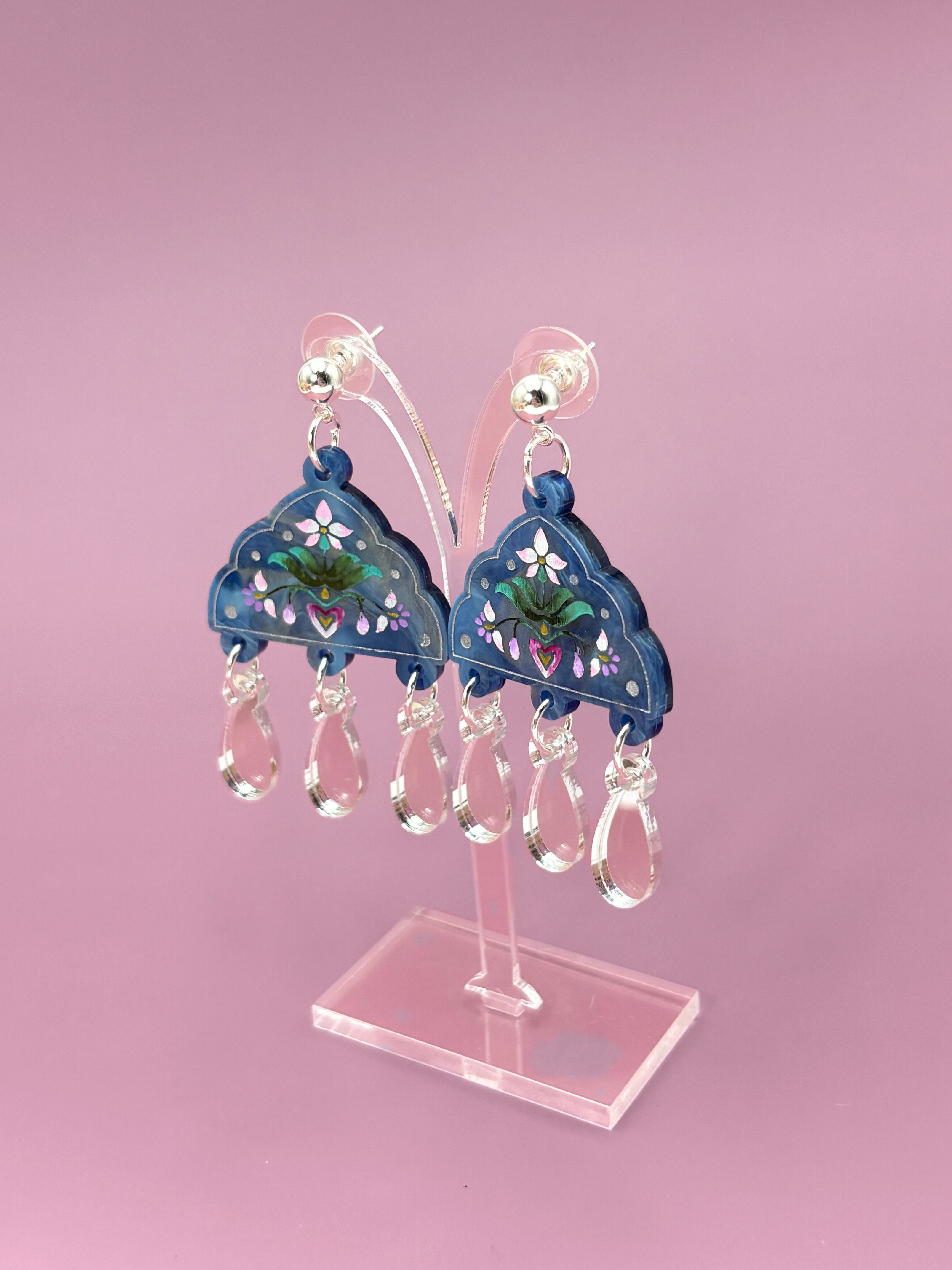 Blue earrings with floral designs on a pink stand against a pink background