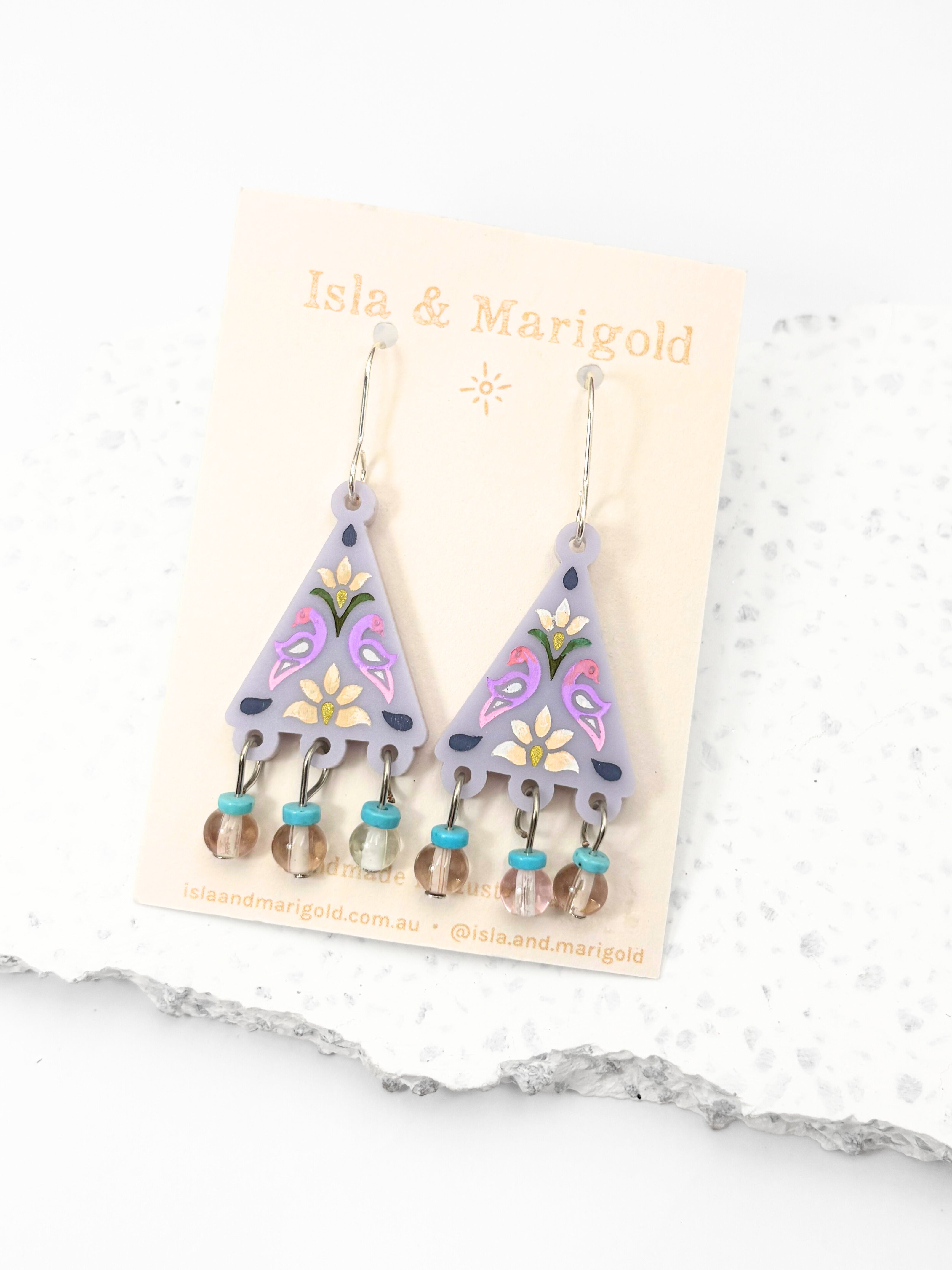 Triangular earrings with floral designs on a 'Isis & Marigold' card against a marble background.