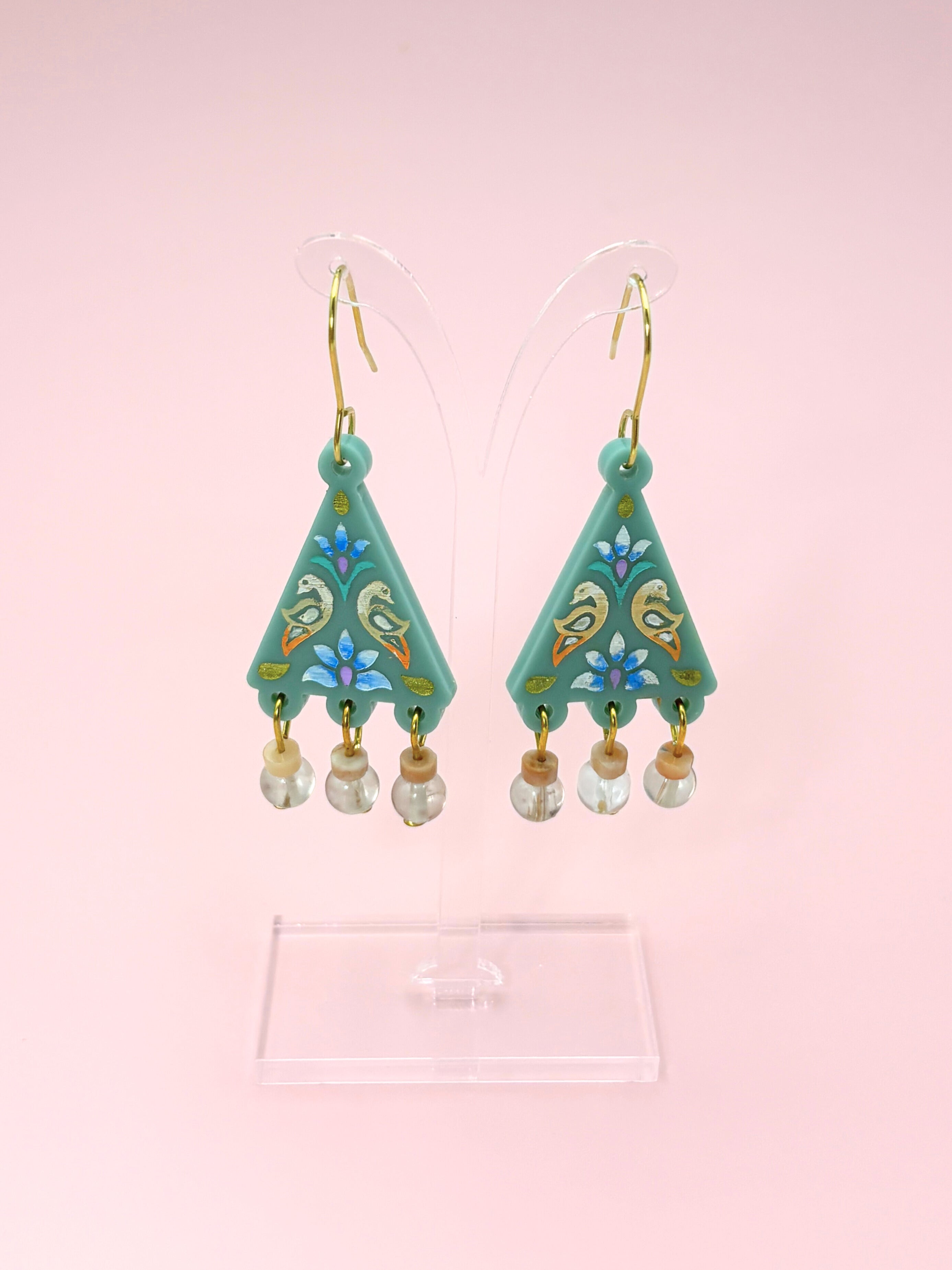 Teal earrings with floral designs and pearl-like beads on a pink background