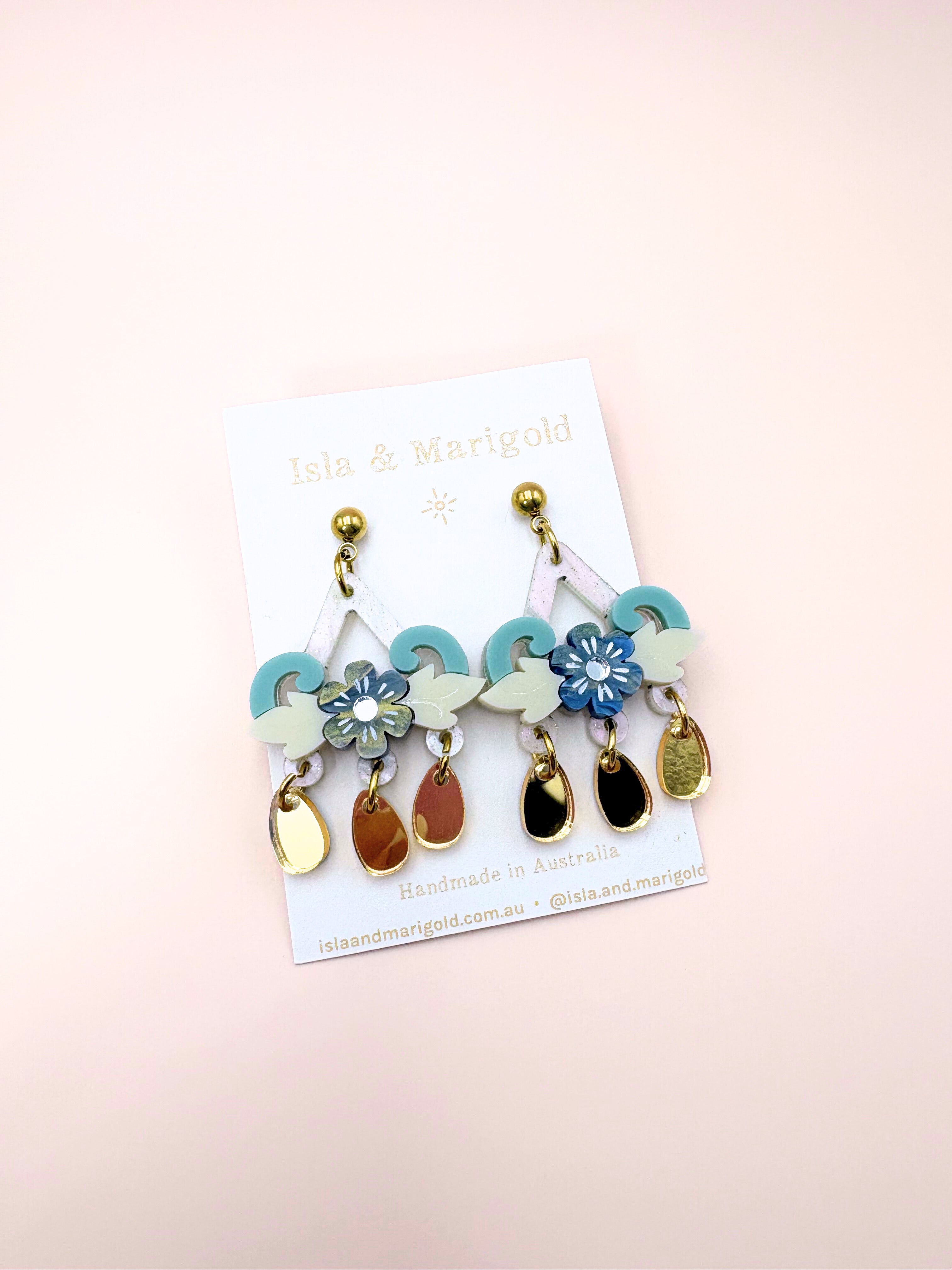 Colorful floral and leaf-shaped earrings on a branded card against a light pink background