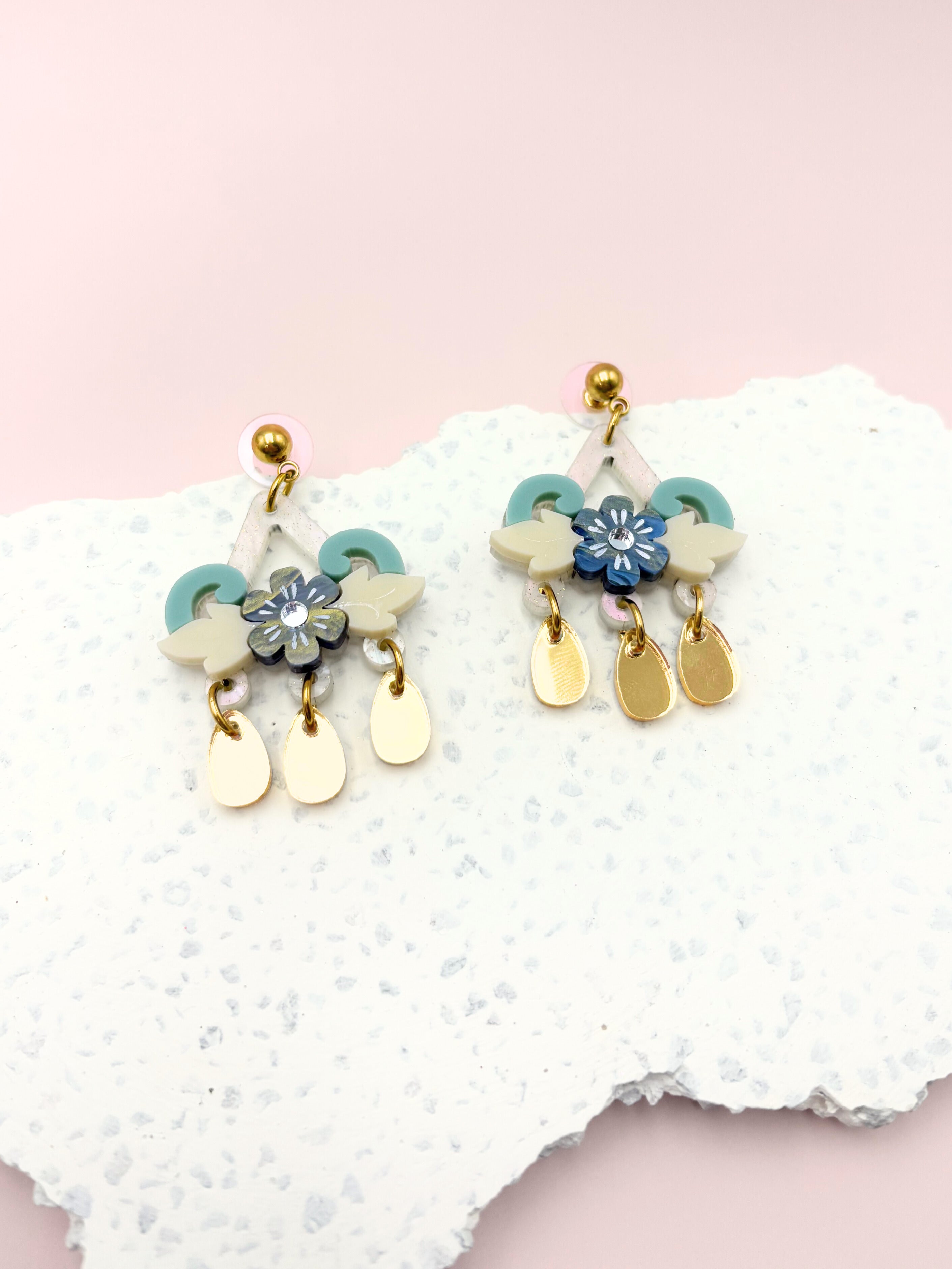 Pair of floral earrings with gold stainless steel and blue accents on a textured white surface. 